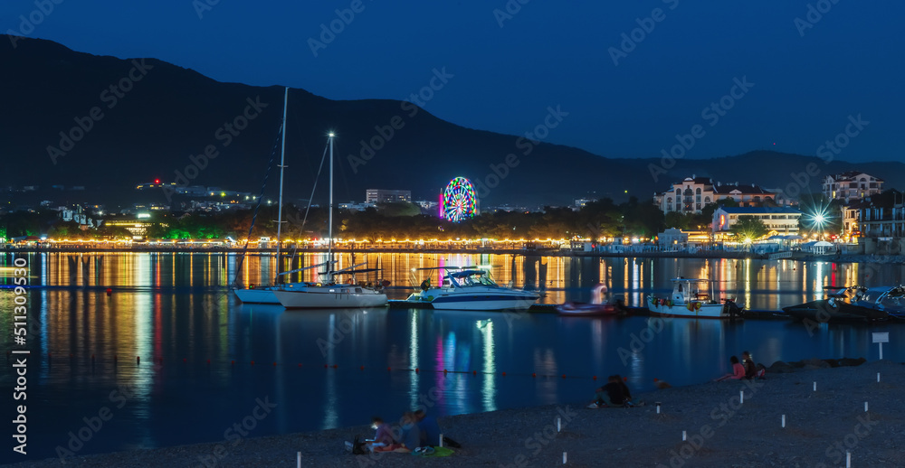 Night photo of sea bay with yachts and boats and reflection of resort town illumination in sea water against backdrop of mountains and blue sky.