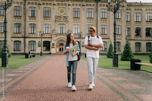 Two international stylish students walk near the campus and smile. Friendly African American guy and Caucasian girl walk together near the university