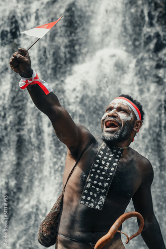 Spirit of Papua man wearing traditional clothes of Dani tribe  red-white headband and bangle is holding little Indonesia flag and celebrating Indonesia independence day against waterfall background. 