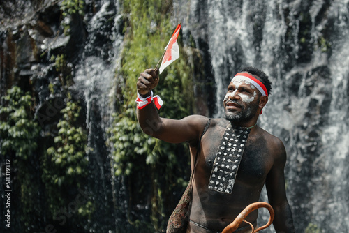 Spirit of Papua man wearing traditional clothes of Dani tribe, red-white headband and bangle is holding little Indonesia flag and celebrating Indonesia independence day against waterfall background.  photo