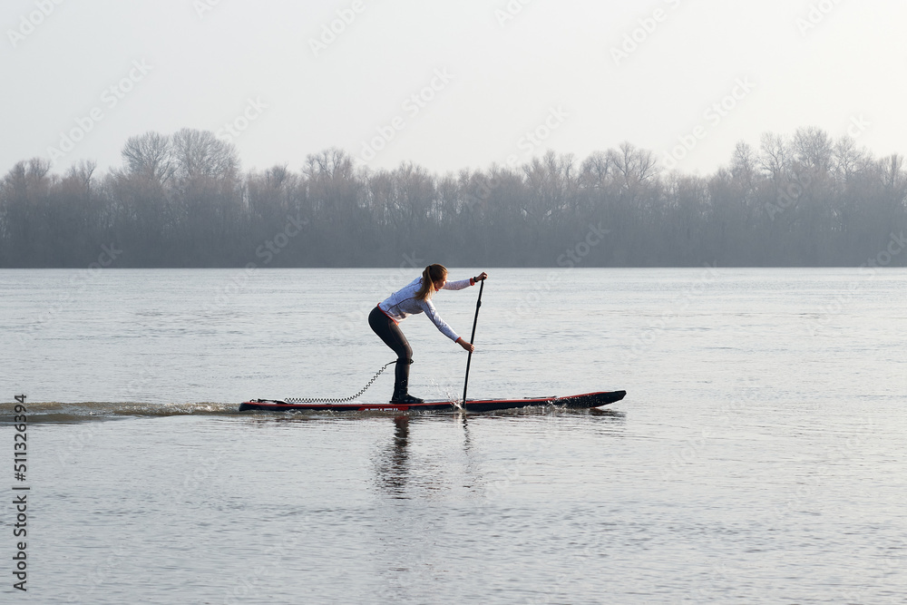 Sporty girl surfing on stand up paddle (SUP) rowing at calm winter Danube river against the background of the shore with trees