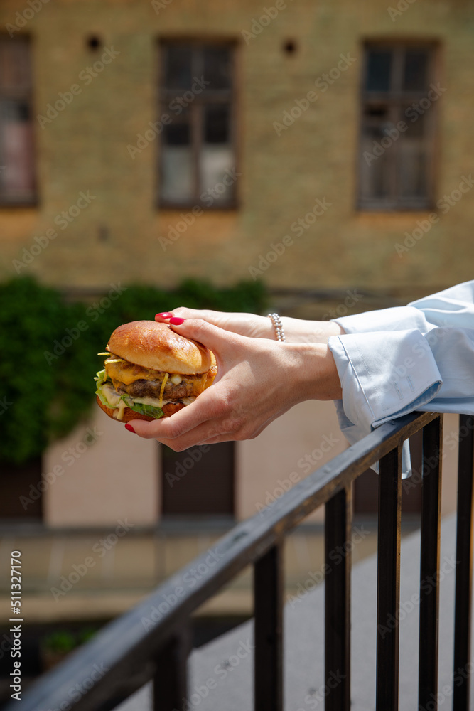 The girl is holding a juicy burger in front of the city. Cheeseburger in women’s hands on the terrace of a fashionable institution. Delicious juicy sandwich. Vertical frame