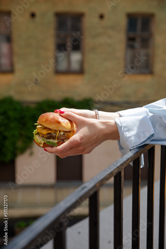 Obraz na płótnie The girl is holding a juicy burger in front of the city. Cheeseburger in women’s hands on the terrace of a fashionable institution. Delicious juicy sandwich. Vertical frame w salonie