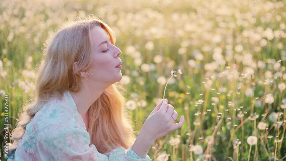 beautiful young blonde woman with long hair blowing on a dandelion in a field at sunset, fabulous video in nature, summer vacation concept.