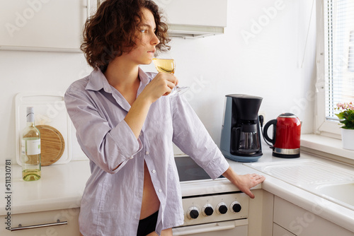 young beautiful romantic woman in men's shirt, buttoned with one button, in bikini, with slim figure, stands alone in kitchen with glass of white wine and looks away