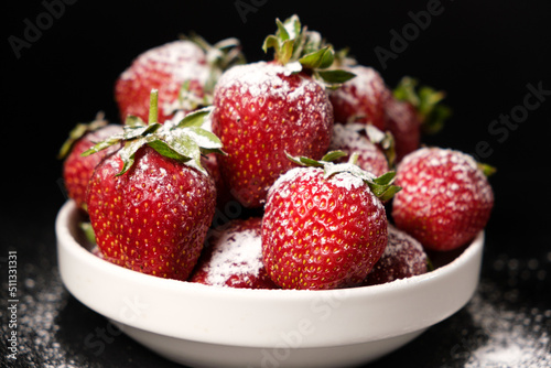 Strawberries in a plate with powdered sugar on a black background, side view close-up.