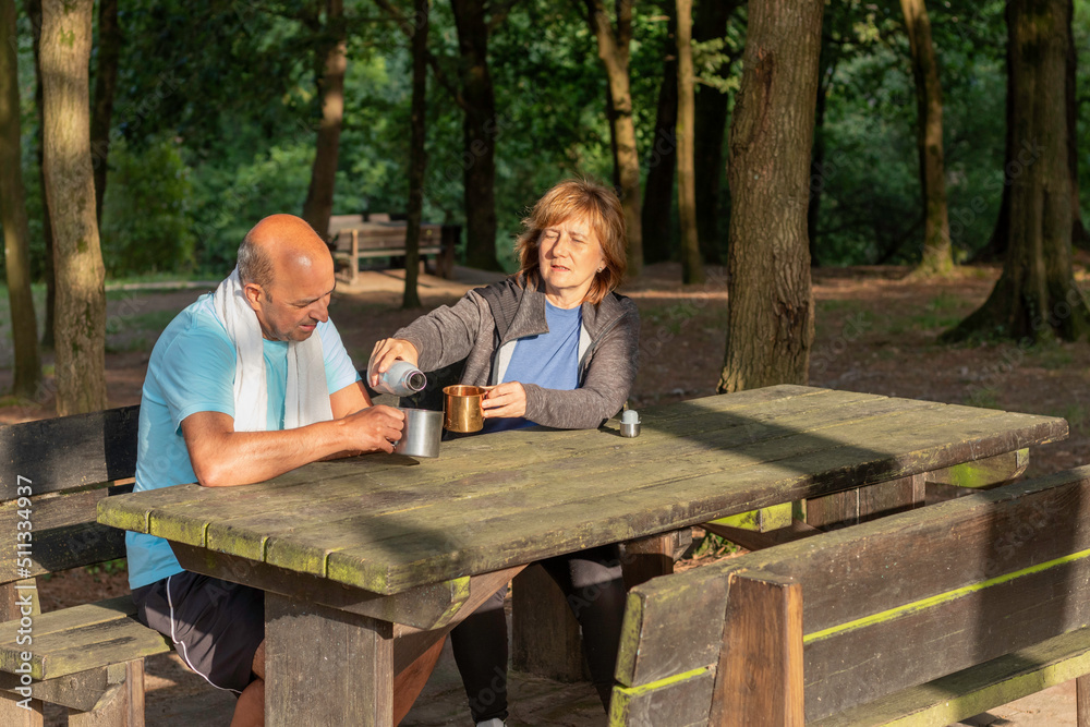 Fototapeta premium Senior couple sitting in a picnic table in the forest drkinking water from bottles in metal camping cups