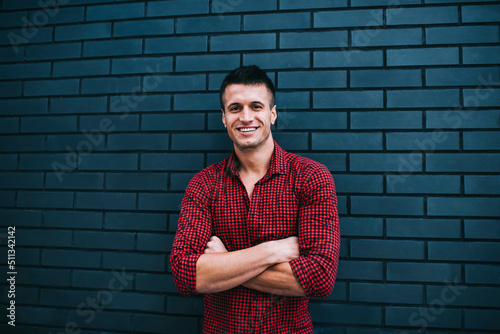 Half length Portrait of 30 years old  happy male in checked shirt standing near copy space wall background. Smiling Handsome young man posing for camera with hands crossed, feeling confidence