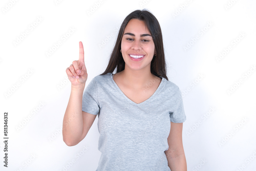 Young beautiful brunette woman wearing grey T-shirt over white wall showing and pointing up with finger number one while smiling confident and happy. Young beautiful brunette woman wearing grey T-shirt over white wall showing and pointing up with finger number one while smiling confident and happy.