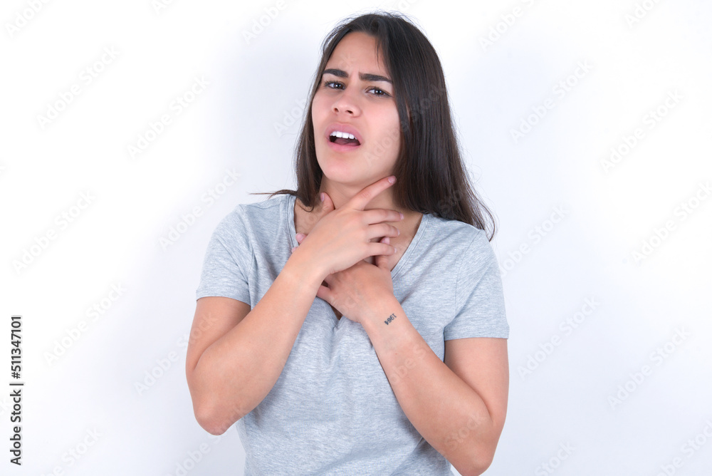 young beautiful brunette woman wearing grey t-shirt over white wall shouting suffocate because painful strangle. Health problem. Asphyxiate and suicide concept.