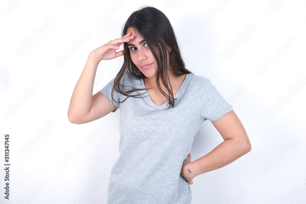 young beautiful brunette woman wearing grey t-shirt over white wall having problems, worried and stressed holds hand on forehead.