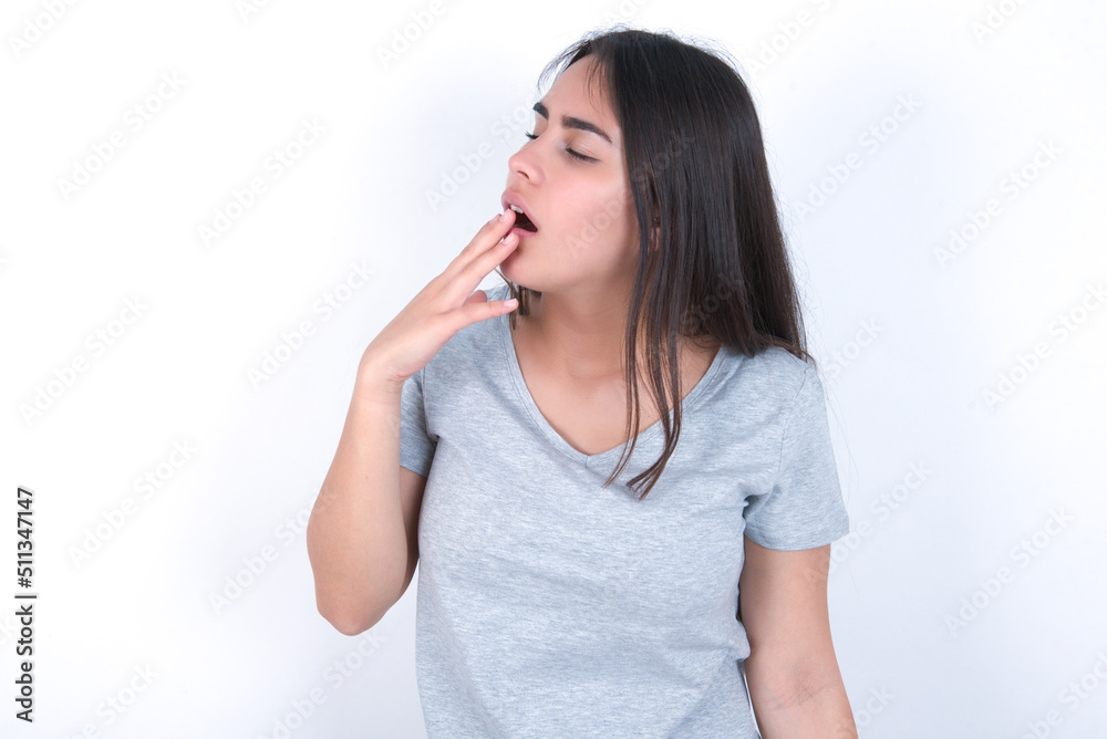 young beautiful brunette woman wearing grey t-shirt over white wall being tired and yawning after spending all day at work. young beautiful brunette woman wearing grey t-shirt over white wall being tired and yawning after spending all day at work.