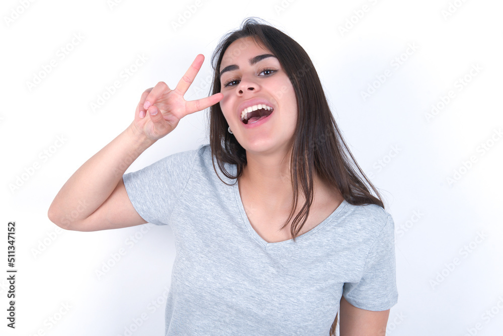 young beautiful brunette woman wearing grey t-shirt over white wall Doing peace symbol with fingers over face, smiling cheerful showing victory young beautiful brunette woman wearing grey t-shirt over white wall Doing peace symbol with fingers over face, smiling cheerful showing victory