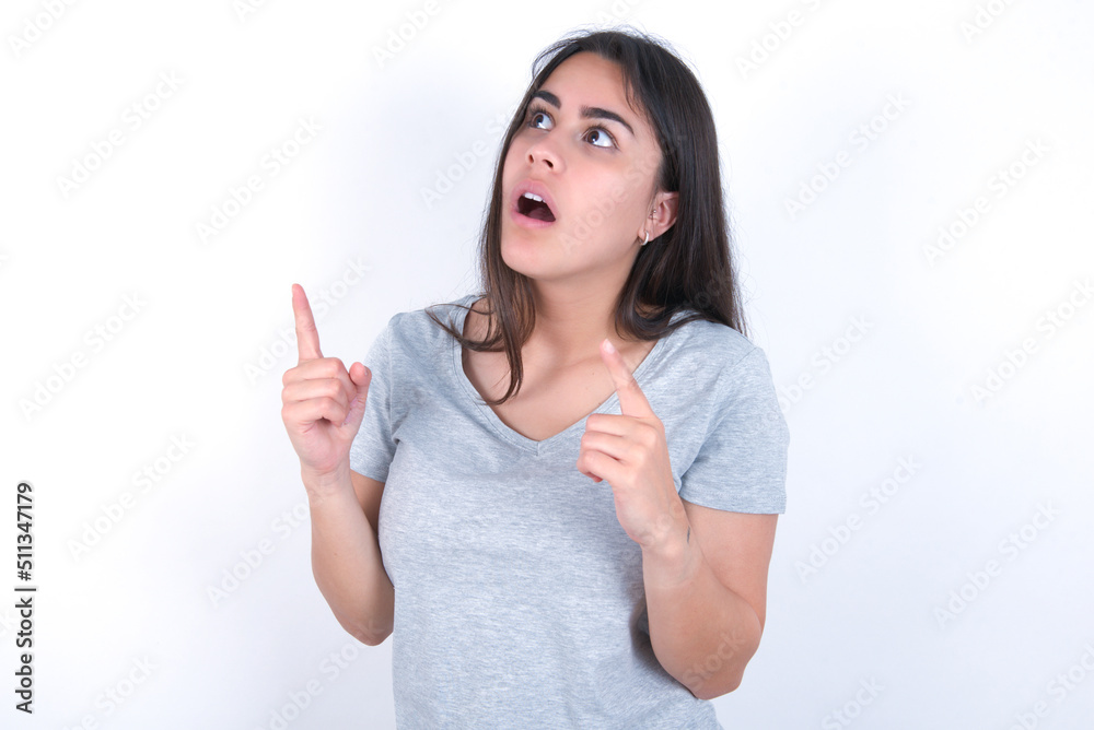 young beautiful brunette woman wearing grey t-shirt over white wall amazed and surprised looking up and pointing with fingers and raised arms. young beautiful brunette woman wearing grey t-shirt over white wall amazed and surprised looking up and pointing with fingers and raised arms.
