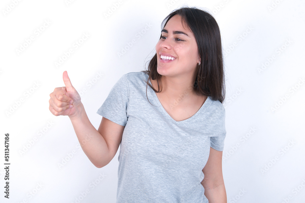 young beautiful brunette woman wearing grey t-shirt over white wall Looking proud, smiling doing thumbs up gesture to the side. Good job! young beautiful brunette woman wearing grey t-shirt over white wall Looking proud, smiling doing thumbs up gesture to the side. Good job!