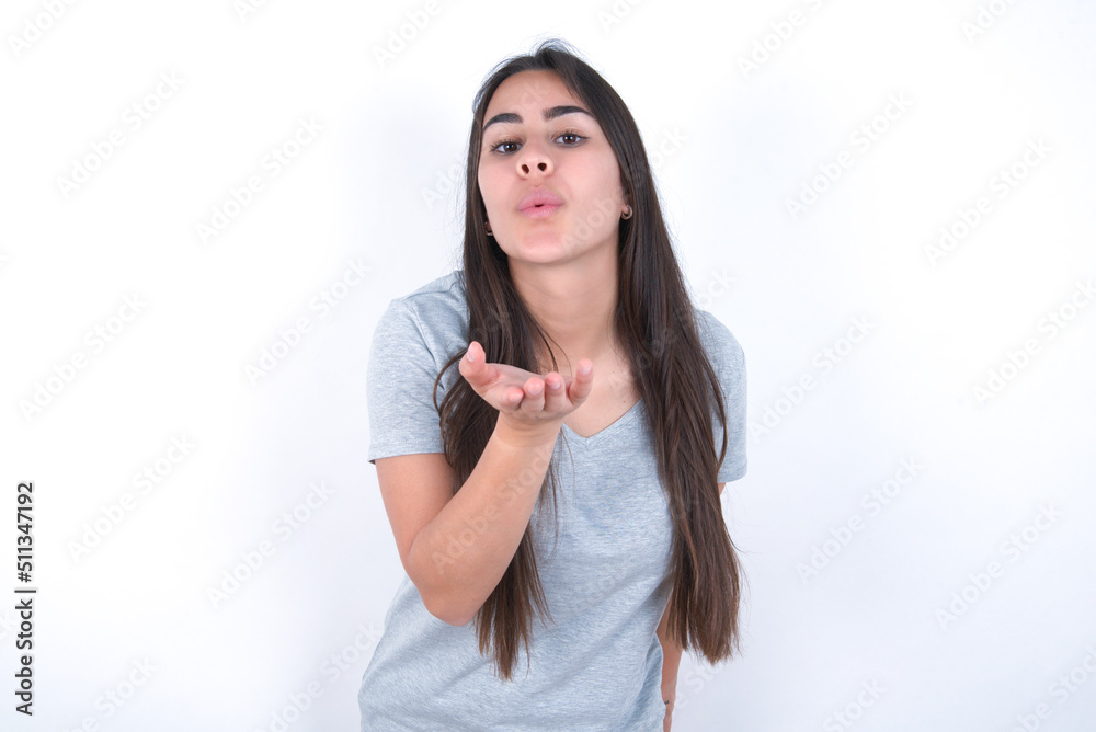 young beautiful brunette woman wearing grey t-shirt over white wall looking at the camera blowing a kiss with hand on air being lovely and sexy. Love expression. young beautiful brunette woman wearing grey t-shirt over white wall looking at the camera blowing a kiss with hand on air being lovely and sexy. Love expression.