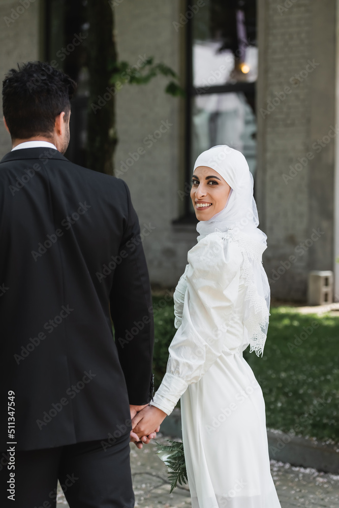 groom holding hands with cheerful muslim bride in wedding dress and walking together.