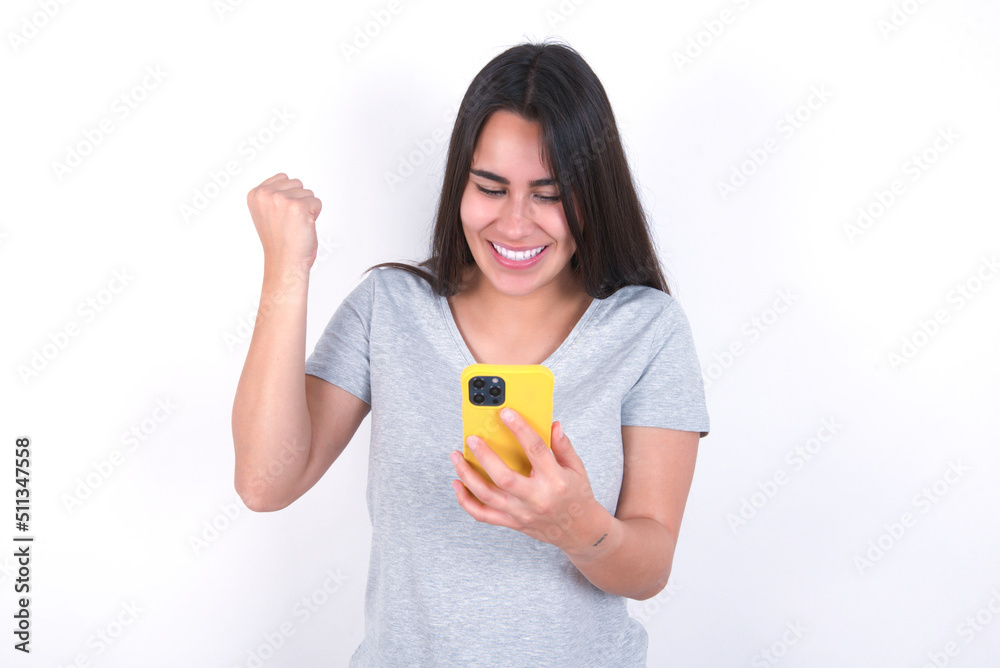 Young beautiful brunette woman wearing gray T-shirt over white wall holding in hands cell and rising his fist up being excited after reading good news.