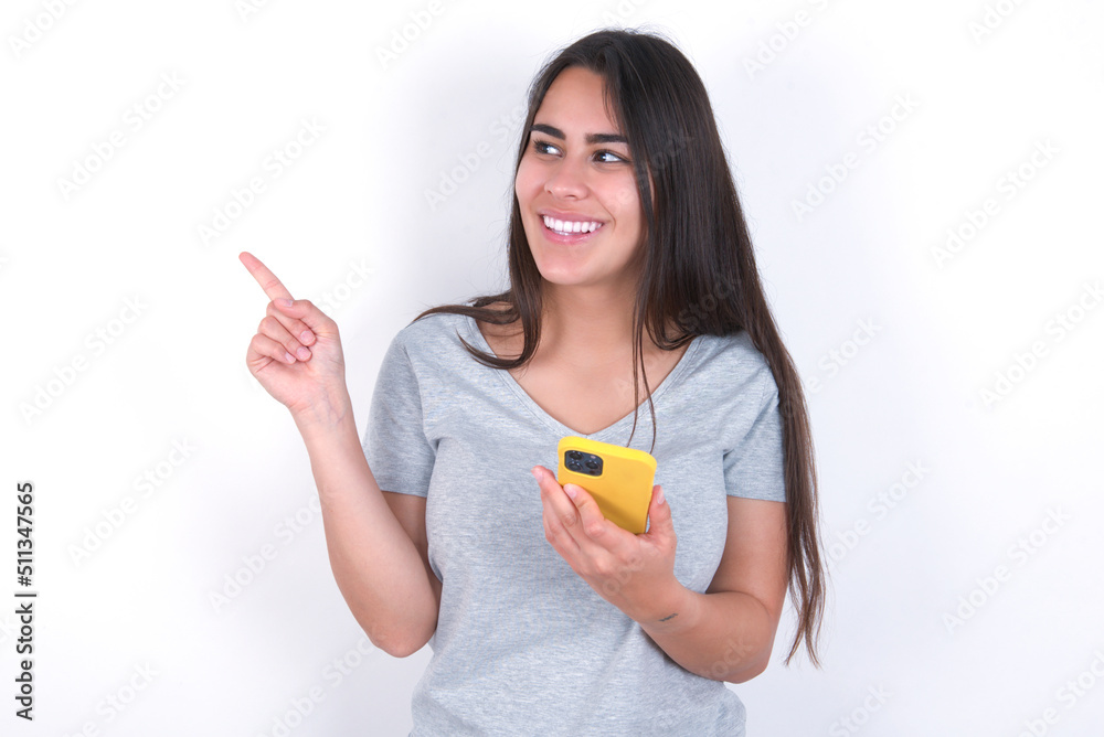 Wow!! excited Young beautiful brunette woman wearing gray T-shirt over white wall showing mobile phone with open hand gesture Wow!! excited Young beautiful brunette woman wearing gray T-shirt over white wall showing mobile phone with open hand gesture