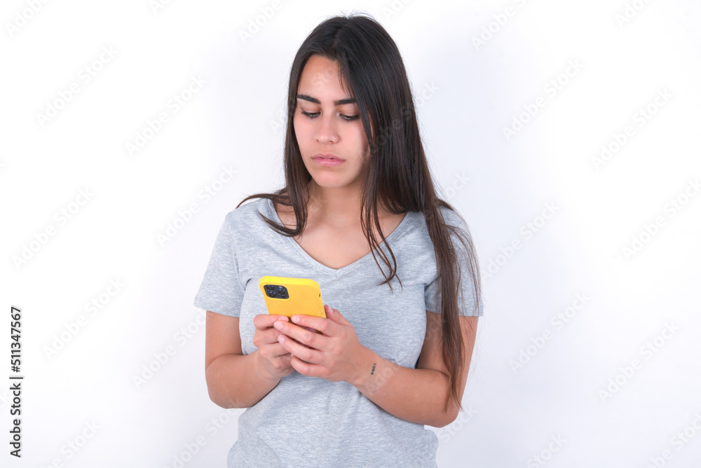 Excited Young beautiful brunette woman wearing gray T-shirt over white wall winking and eye hold smart phone use read social network news Excited Young beautiful brunette woman wearing gray T-shirt over white wall winking and eye hold smart phone use read social network news