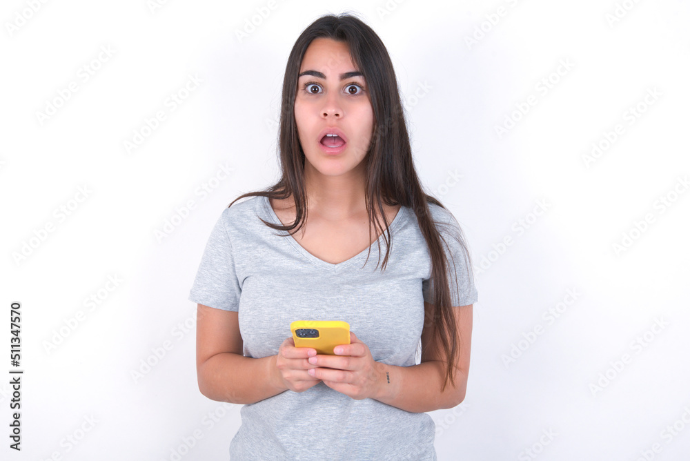 Shocked Young beautiful brunette woman wearing gray T-shirt over white wall opens mouth hold phone reading advert unbelievable big shopping prices Shocked Young beautiful brunette woman wearing gray T-shirt over white wall opens mouth hold phone reading advert unbelievable big shopping prices