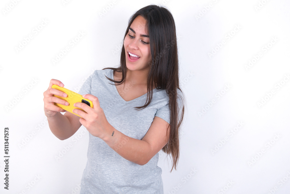 Portrait of an excited Young beautiful brunette woman wearing gray T-shirt over white wall playing games on mobile phone. Portrait of an excited Young beautiful brunette woman wearing gray T-shirt over white wall playing games on mobile phone.