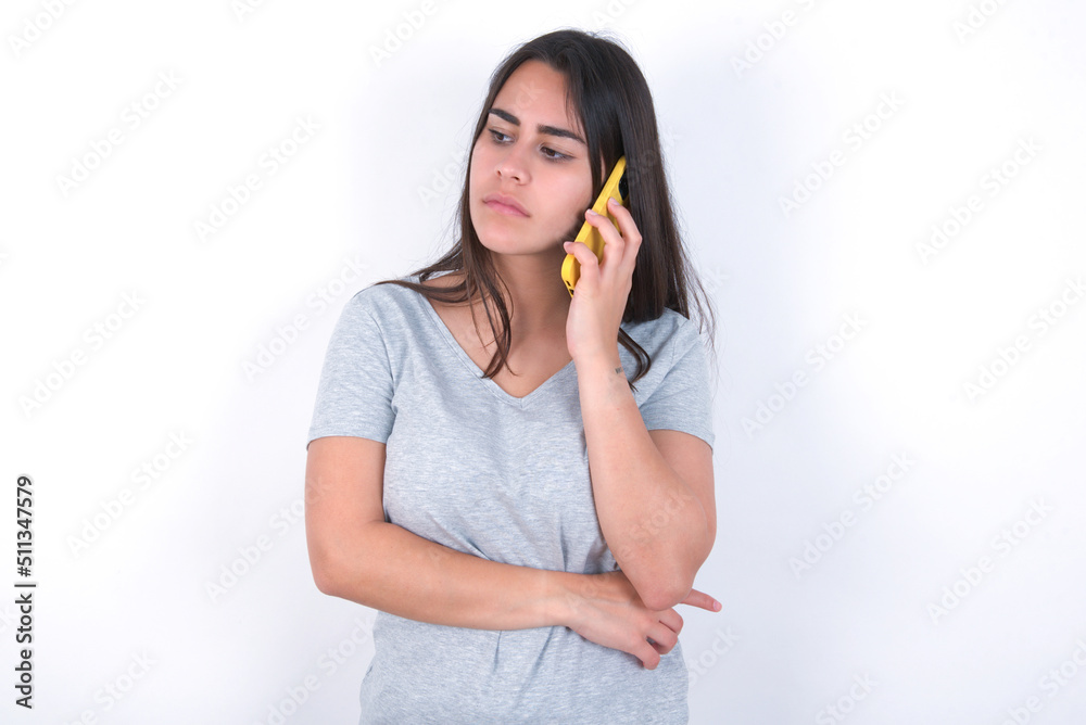 Sad Young beautiful brunette woman wearing gray T-shirt over white wall talking on smartphone. Communication concept. Sad Young beautiful brunette woman wearing gray T-shirt over white wall talking on smartphone. Communication concept.