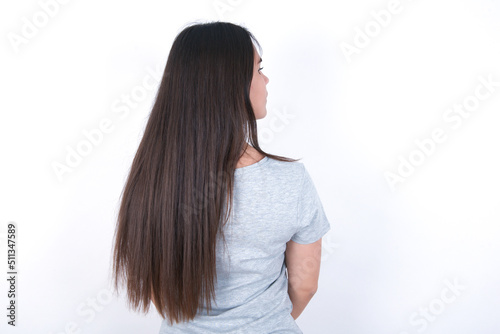 The back side view of a Young beautiful brunette woman wearing gray T-shirt over white wall. Studio Shoot.