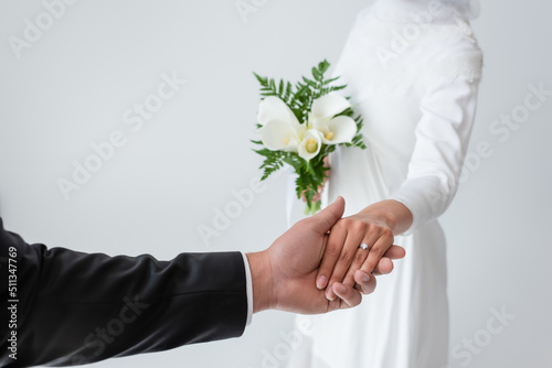 Print op canvas cropped view of bride with bouquet holding hands with blurred groom isolated on grey