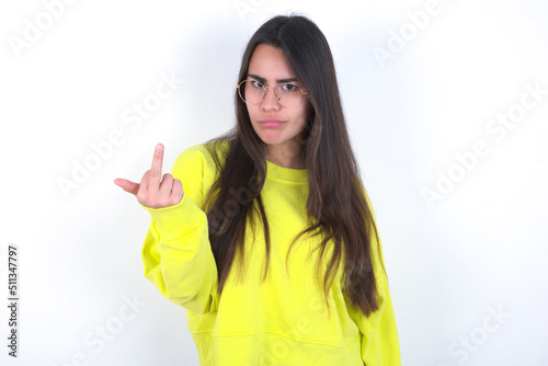 Young beautiful brunette woman wearing yellow hoodie over white wall shows middle finger bad sign asks not to bother. Provocation and rude attitude. photo