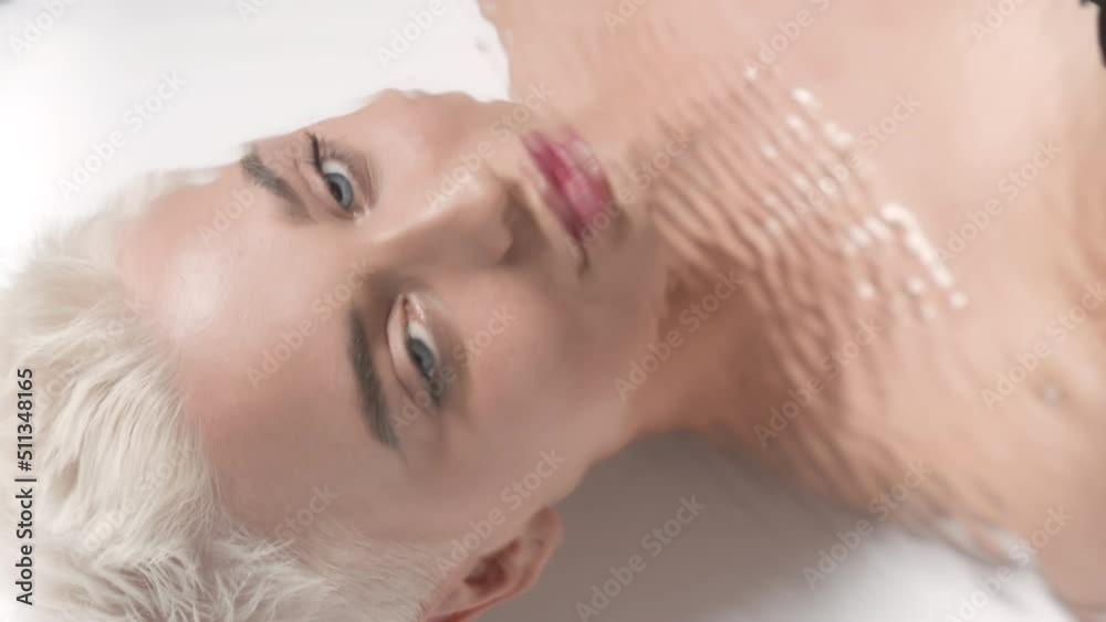 Pretty young slim Caucasian woman with short white hair turns her head to the camera and opens her eyes looking at it through rippling water on white background | Droopy eyes prevention concept Pretty young slim Caucasian woman with short white hair turns her head to the camera and opens her eyes looking at it through rippling water on white background | Droopy eyes prevention concept