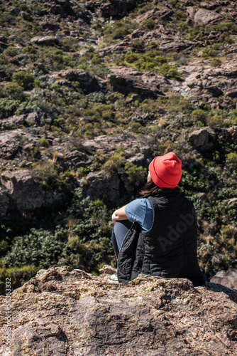 Young woman with a red hat sitting on the rocks looking out from a mountain in Cordoba