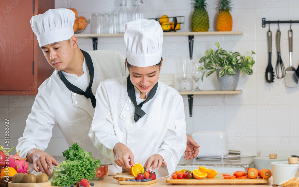 Two Asian professional couple chef wearing white uniform and hat, helping for preparing ingredients for healthy meal, cooking in kitchen together, smiling with happiness and confidence. Food Concept.