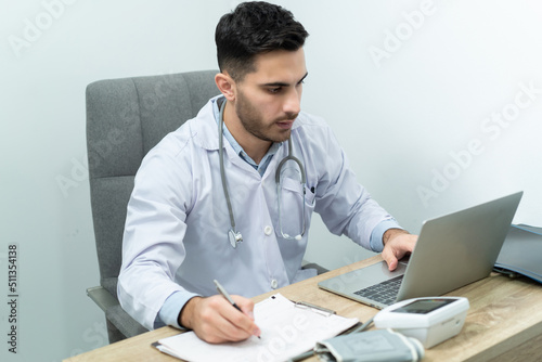 Young Caucasian male doctor in white medical gown with a stethoscope around his neck sitting at a working desk, working with laptop and writing notes on a medical clipboard in clinic examination room.