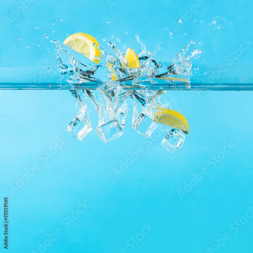 Water splash with fresh lemon fruit slices and ice cubes. Minimal summer vacation pool or beach cocktail drink party concept. Fun bright blue background.