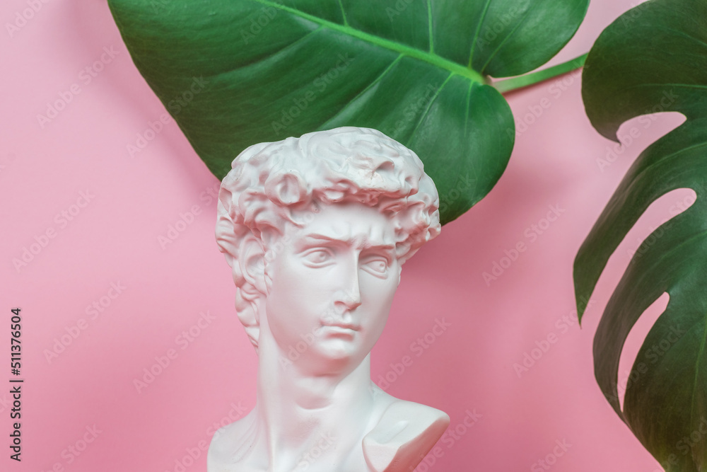 An antique statue on a pink background with large green leaves. Minimal concept.