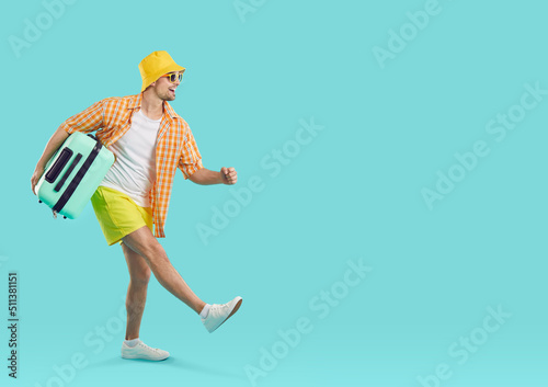 Happy excited guy tourist or vacationer, goes carrying travel suitcase on light blue background. Joyful young man in summer clothes and with suitcase in hand walks towards copy space. Full length. photo
