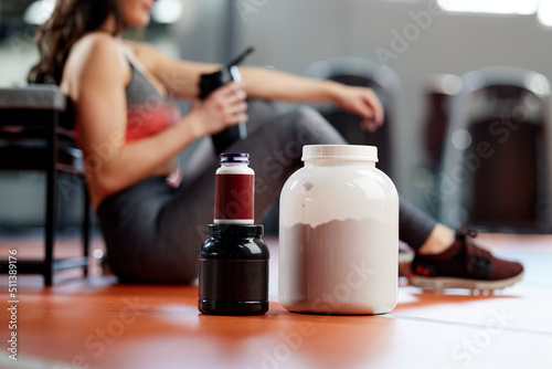 Selective focus on protein powder and multivitamins jars on the gym floor with a sporty woman in background.