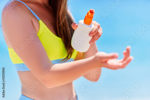 Female applying and holding white empty bottle blank of sunscreen spray lotion while sunbathing on beach by the sea in sunny summer day. Sun protection