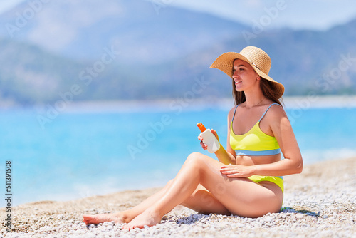 Young girl applying sunscreen lotion spray while sunbathing and relaxing on beach by the sea in sunny summer day. Sun protection