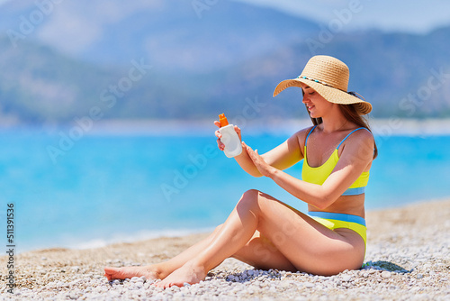 Young girl applying sunscreen spray lotion while sunbathing and relaxing on beach by the sea in sunny summer day. Sun protection