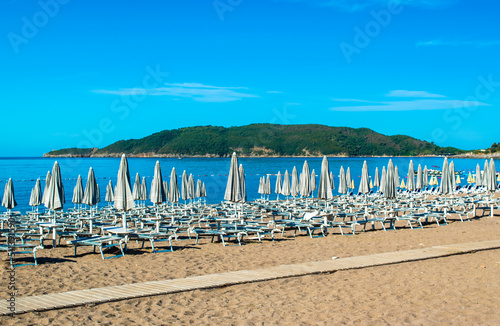 Sun loungers and umbrellas on public beach at summer sunny mornin. Skojl island background. Calm sea and blue sky Becici  Montenegro.