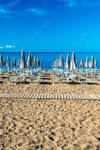 Sun loungers and umbrellas on public beach at summer sunny mornin. Skojl island background. Calm sea and blue sky Becici, Montenegro. © jana_janina