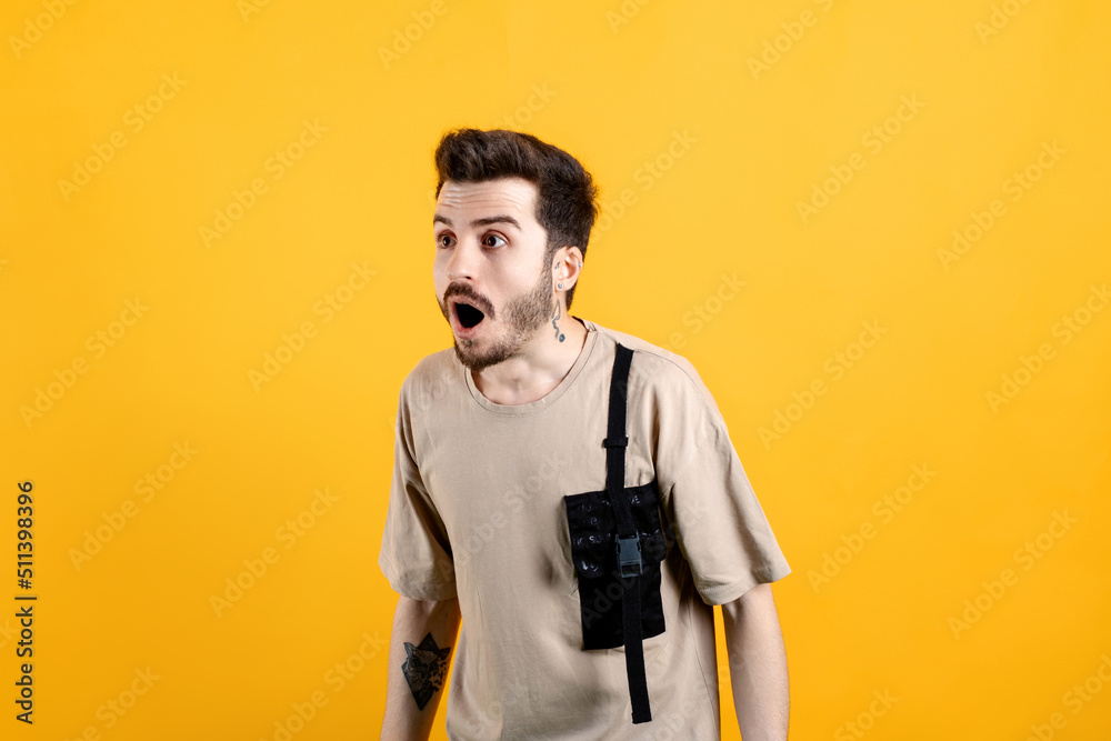 Young man wearing beige t-shirt posing isolated over yellow background looking left with big eyes and open mouth, shocked by news, empty copy space. Young man wearing beige t-shirt posing isolated over yellow background looking left with big eyes and open mouth, shocked by news, empty copy space.