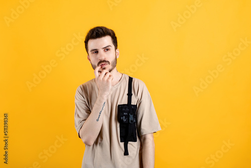 Handsome young man wearing casual clothes posing isolated over yellow background looking at upper right corner touching lip, thinking. Trying think up solution.