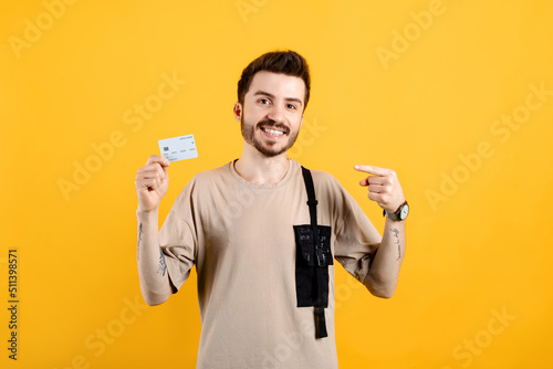 Cheerful young man wearing t-shirt posing isolated over yellow background pointing finger at credit or debit card. Shopping and finance concept. © platinumArt