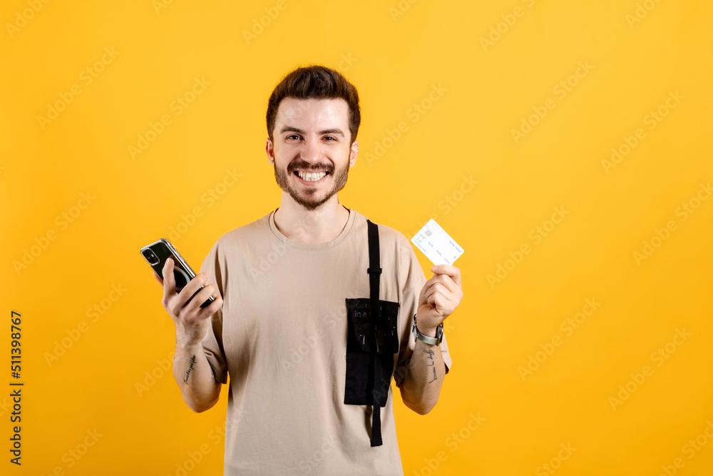 Happy casual man wearing t-shirt posing isolated over yellow background holding credit card and mobile phone while smiling and looking at the camera. Happy casual man wearing t-shirt posing isolated over yellow background holding credit card and mobile phone while smiling and looking at the camera.