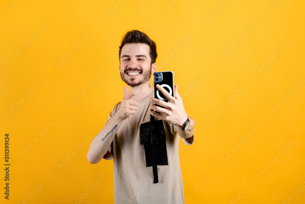 Happy young man wearing beige t-shirt posing isolated over yellow background pointing index finger on mobile phone while taking selfie with big smiles. Communicating concepts. Happy young man wearing beige t-shirt posing isolated over yellow background pointing index finger on mobile phone while taking selfie with big smiles. Communicating concepts.