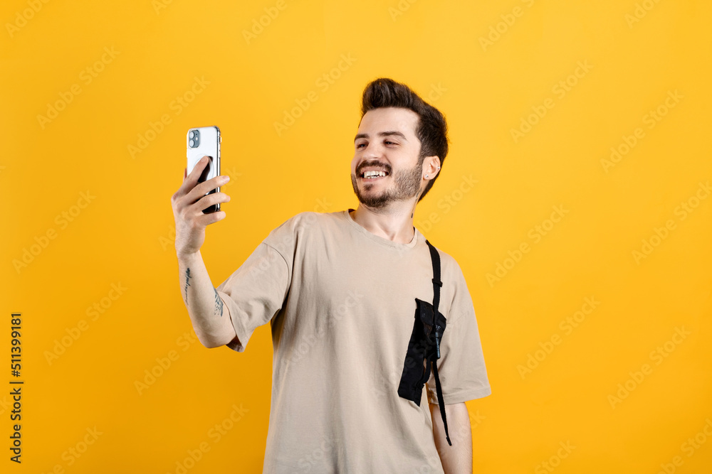 Young man wearing beige t-shirt posing isolated over yellow background making a selfie looking at the phone screen with big smiles. Young man wearing beige t-shirt posing isolated over yellow background making a selfie looking at the phone screen with big smiles.