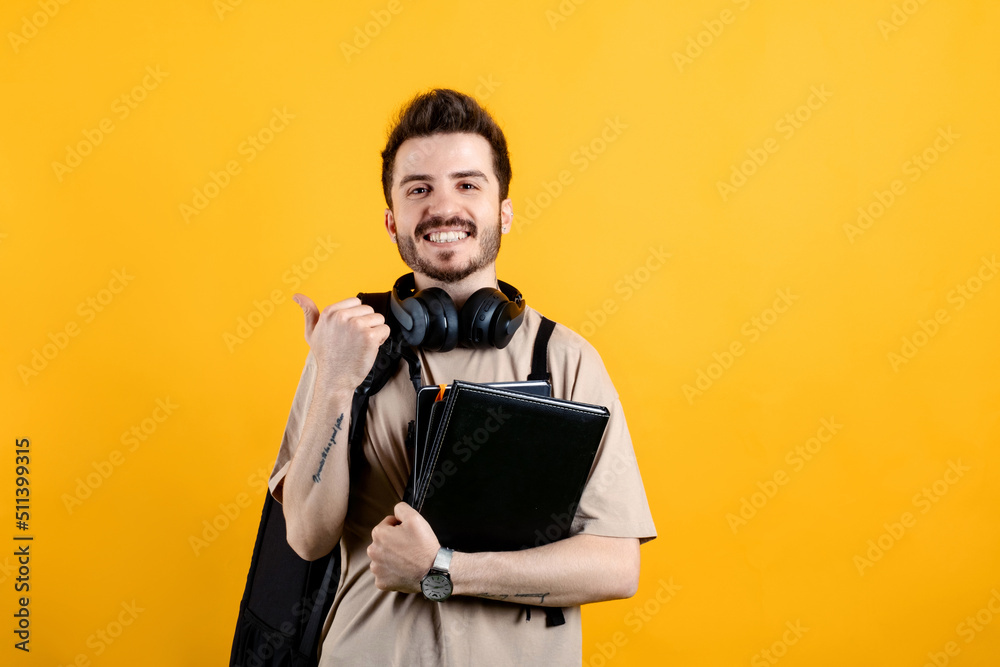 Handsome caucasian man wearing beige t-shirt posing isolated over yellow background smiling with happy face looking and pointing to the side with thumb up. Handsome caucasian man wearing beige t-shirt posing isolated over yellow background smiling with happy face looking and pointing to the side with thumb up.